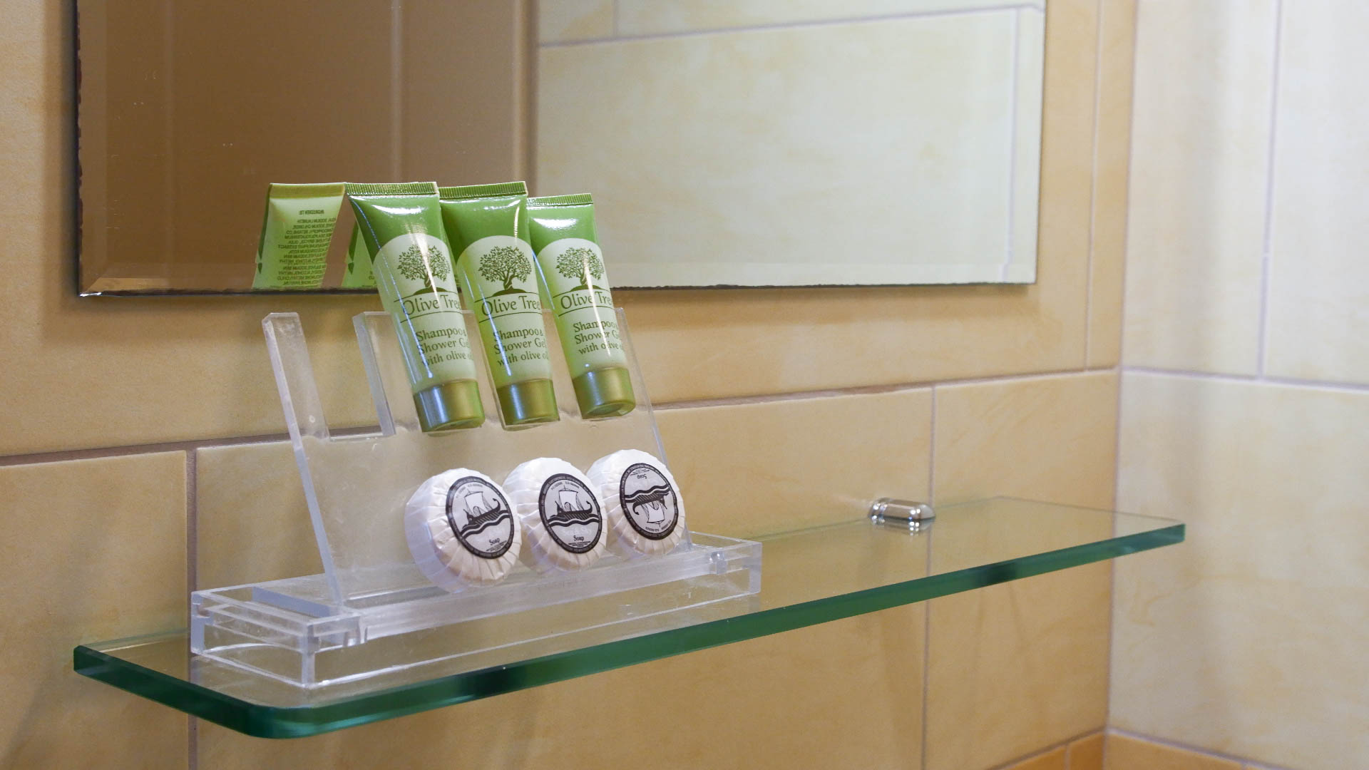 Three small shampoos and soaps on a glass surface in front of the mirror.